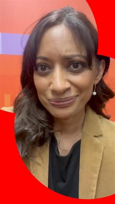 Dharshini David has long brown hair and wears a camel jacket and black shirt