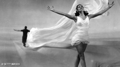 Cyd Charisse wears a white dress, with her arms up in the air