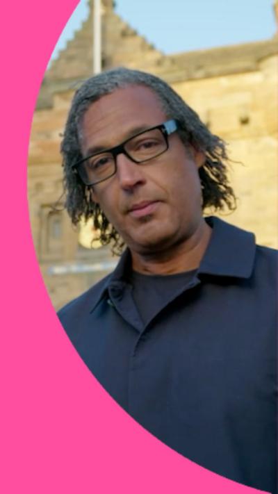 A man, Professor David Olusoga, with long dark hair, glasses and wearing a blue top.