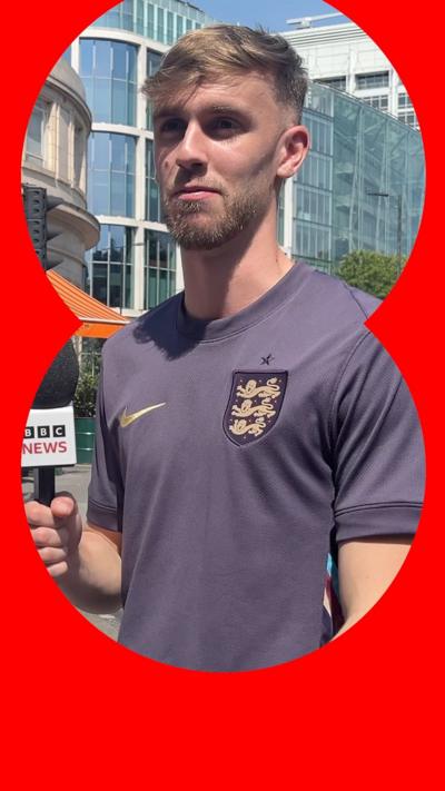 A man wearing a purple top, holding a BBC News microphone.