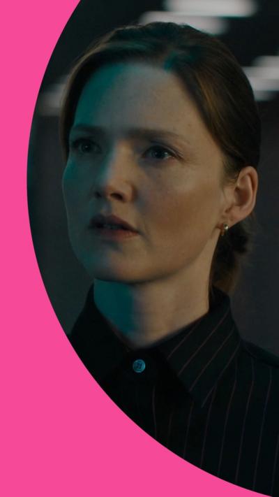 A woman with dark hair pulled back from her face looks at someone off camera, she wears a dark shirt