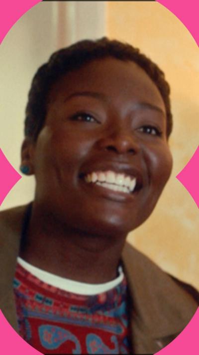 A woman smiles in a living room. She wears a brown jacket and a colourful top underneath