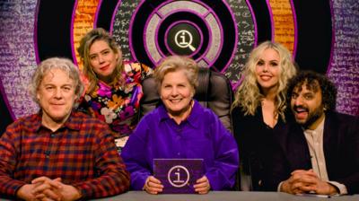 Five people, three women and two men, sit behind a desk together. The QI logo can be seen behind them.