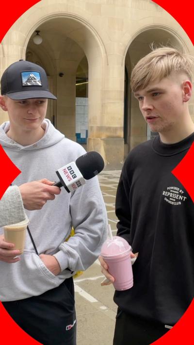 Two young men are being interviewed. One wears a grey hoodie and black hat, the other wears a black sweater