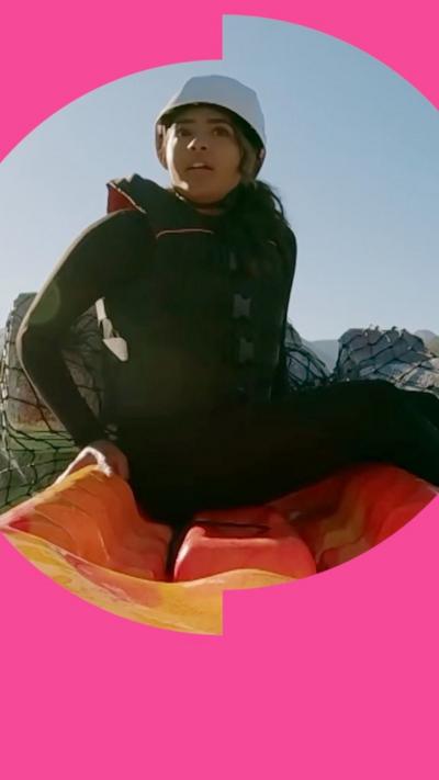 A woman wearing a wetsuit and white helmet sits on an orange kayak