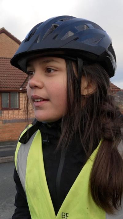 A young girl with long dark hair stood at the side of a road with a black cycle helmet and green hi vis jacket