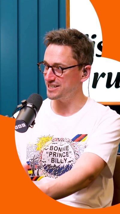 Man with short brown hair and glasses, wears a white tshirt and sits in front of a microphone