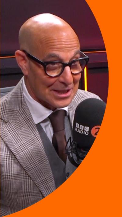 Stanley Tucci is wearing a grey suit with a brown tie and glasses. He is sitting in front of a BBC Radio 2 microphone.