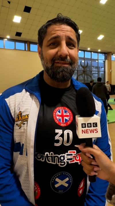 Man wearing kabaddi uniform speaking into BBC microphone, standing in indoor sports hall