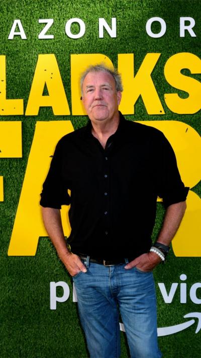 Jeremy Clarkson stands in front of a green wall with the words Clarkson's Farm written on it in yellow. He is wearing a black shirt with the sleeves rolled up and a pair of blue jeans. He has his hands in his pockets. 