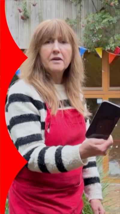 A woman outside with red apron holding a black phone.