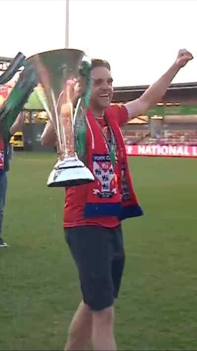 York City footballer in red team shirt punches the air as he carries a trophy around the picth during a promotion celebration with fans