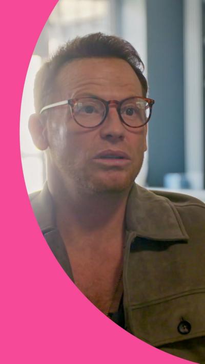 Joe Swash has short dark red hair and wears brown circular glasses and a khaki jacket