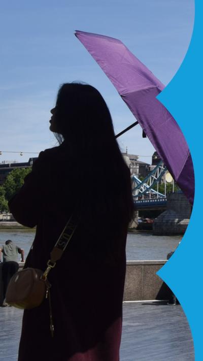 A woman stands silhouetted on the banks of the River Thames, holding a purple umbrella for shade.