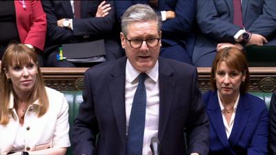 Prime Minister Sir Keir Starmer, wears a blue suit and glasses, as he stands in the House of Commons. Angela Rayner and Rachel Reeves sit either side of him on the benches.