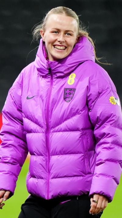 England goalkeeper Hannah Hampton smiles on the pitch. She is wearing a pink puffer jacket.