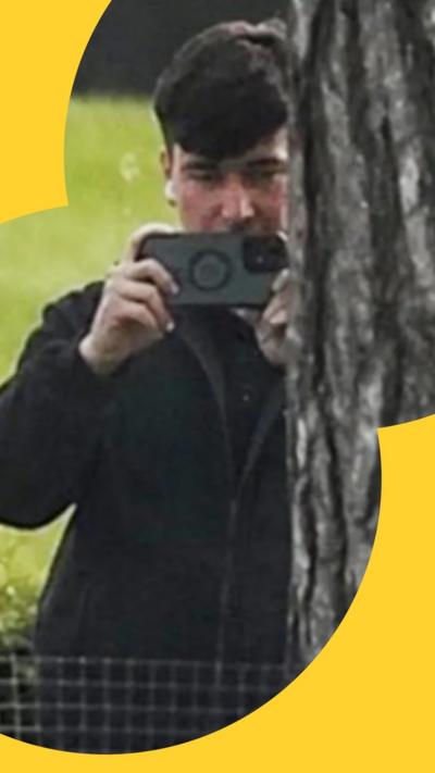 A man with short dark hair stands next a tree filming with his phone