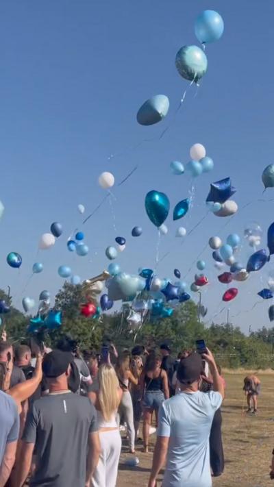 A crowd of people stood on grass against the backdrop of a blue sky with arms outstretched to release the balloons.