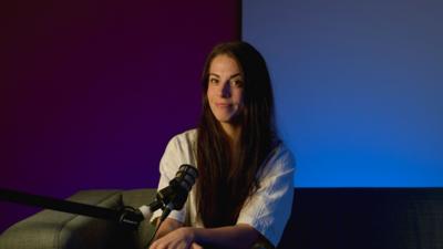A young woman with long, dark hair and a white top sitting in a dark radio studio with a microphone.