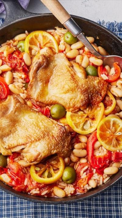  A round pan of Mediterranean chicken with tomatoes, beans, rice, olives and lemon slices is in the middle of the image. It is sitting on a table on top of a blue and white check tablecloth. There is a pile of light blue plates on the left hand side with silver cutlery placed on top.