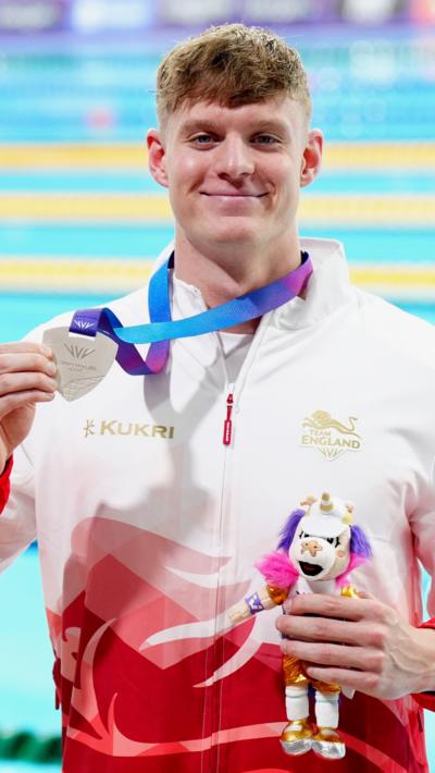 A swimmer in a white and red tracksuit top holding a silver medal attached to a purple ribbon. He's also holding a toy mascot