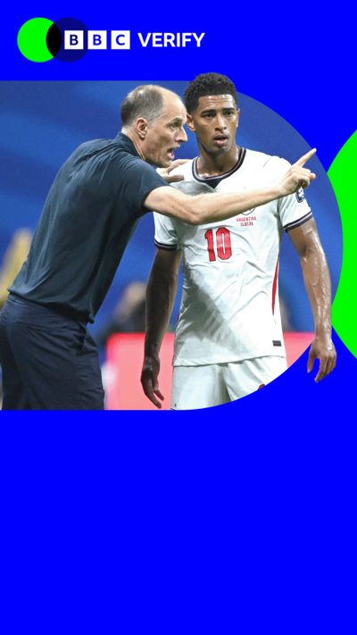 Jude Bellingham wears a white football kit and stands on a pitch next to a referee who is pointing a finger 