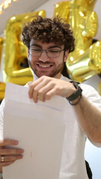 A young man with glasses and brown curly hair opens an envelope to get his A-level results.