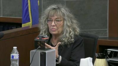 A woman with curly grey hair sits in a black chair in court.