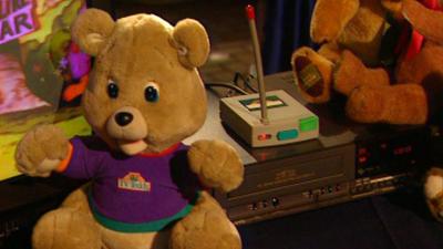 The TV teddy, wearing a purple t-shirt, sits beside a VCR with other teddies on top of it.