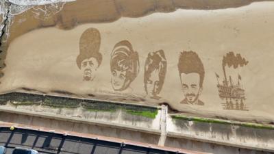 An aerial view of a sandy beach with drawings of Winston Churchill, Princess Diana, John Lennon, David Beckham and a steam train