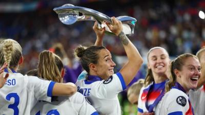 England player Lucy Bronze smiling and holding up the European Championship trophy, surrounded by fellow players. Lucy and her fellow England players are all smiling after their victory