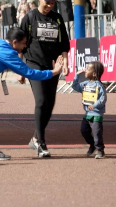 Child giving a high-five to a crouching man in a blue New Balance jacket during a race event finish line.