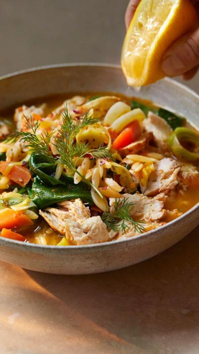 A bowl of lemon chicken orzo soup with a hand squeezing a lemon into it