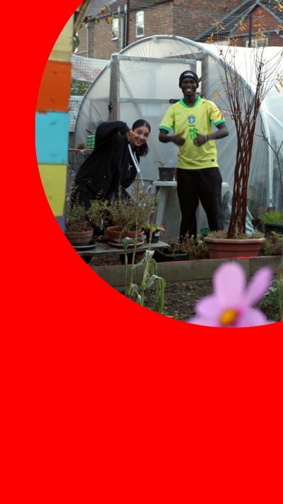 A young woman and a young man smile and make poses in a garden, standing in front of a greenhouse