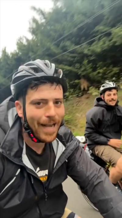 Two men on bikes. They are wearing coats and helmets. There are trees in the background. 
