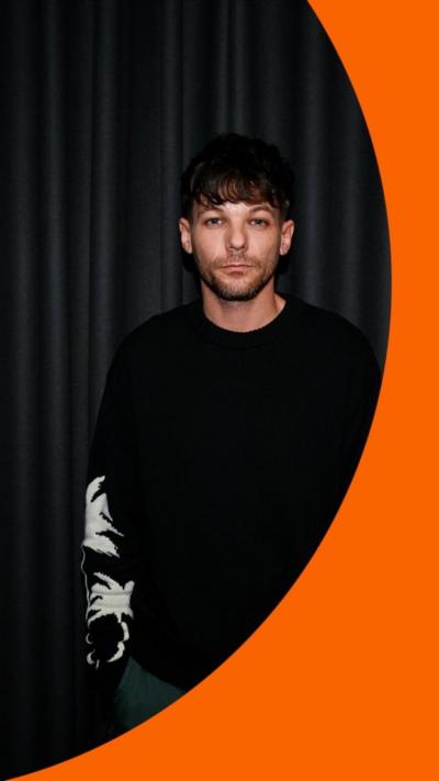 A man, Louis Tomlinson, stands in a black jumper against a dark curtain