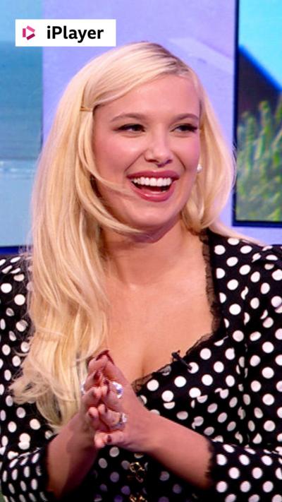 Millie Bobby Brown with long blonde hair, wearing a white and black top.