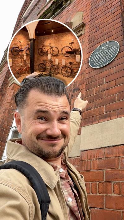 The image shows Josh Gorroño Chapman pointing at a commemorative plaque on the exterior of a historic red-brick building with arched windows. An inset reveals an indoor display of vintage bicycles mounted on a brick wall.
