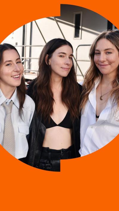 Three members of Haim pose together. They all have long brown hair. Two wear white shirts, one in a black jacket over a bikini top