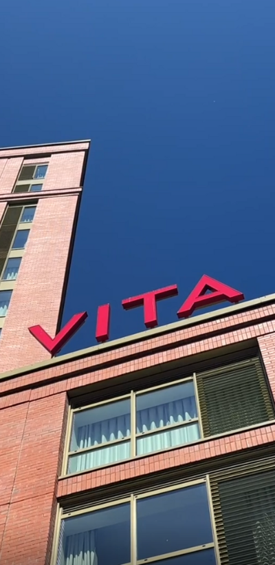 A red brick building in taken from below so the blue sky is visible overhead. It hasa red sign saying VITA on the ro