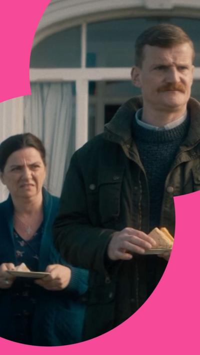 A woman looks in disgust at a moustached man who is in front of her. He is holding a plate of sandwiches