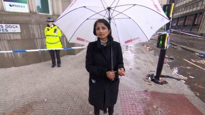 News correspondent Rajini Vaidyanathan holding an umbrella and stood on a street in front of police cordon tape