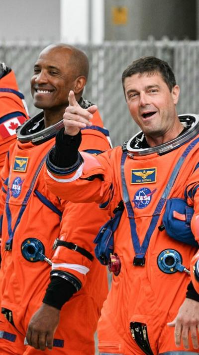 Astronauts wearing orange space suits smile and point to a crowd of well-wishers