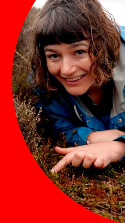 A woman with short brown hair, in a blue waterproof coat, lies down and points at some moss
