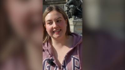 Blonde woman wearing a pink jacket looks to the side with an awkward smile.