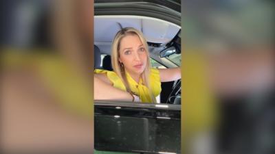 Presenter Emma Vardy looks out of the window from the driving seat of a car. She has long blonde hair and wears a yellow top