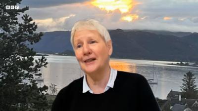 Magaidh Choinneagan, Cathraiche A' Chomuinn Ghàidhealaich, agus i na suidhe le dealbh den a' mhuir agus na beanntan air a cùlaibh. 