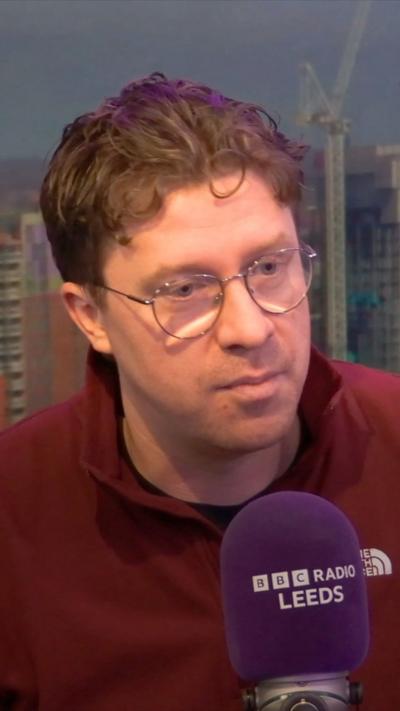 Man in red fleecy sweater wearing spectacles sits in front of a purple microphone branded with BBC Radio Leeds.