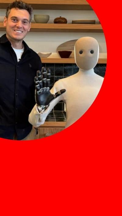 A man, Joe Tidy, wears a black sweater while standing next to a humanoid robot