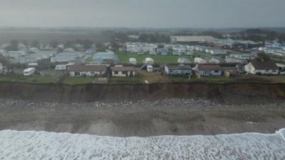 Holiday chalets and caravans are close to the edge of a cliff-top. Below them is a sandy beach with white stones and the tide coming in, which is foamy and white.